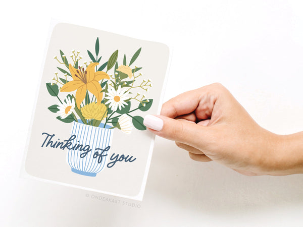 A hand holds the Onderkast Studio Thinking of You Flowers Greeting Card, featuring a striped vase with yellow and white flowers. The card says Thinking of you in script—an elegant floral design for any occasion.