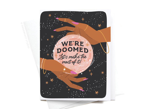The We’re Doomed Greeting Card by Onderkast Studio features two hands with pink nails, gold jewelry, and a glowing crystal ball reading WERE DOOMED Lets make the most of it! on a starry black background—perfect dark humor.