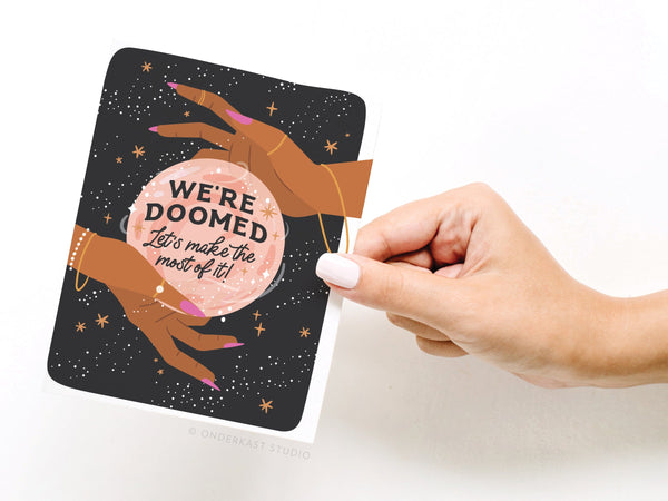 A hand holds Onderkast Studio’s We’re Doomed Greeting Card, featuring two hands around a crystal ball that says “WE’RE DOOMED. Let’s make the most of it!” Stars and sparkles highlight the darkly humorous design.