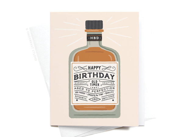 The Onderkast Studio Happy Birthday Whiskey Bottle Greeting Card features a bottle labeled “Happy Birthday” with phrases like “Old Timer,” “Aged to Perfection,” and “Authentic,” shown resting on a crisp white envelope.