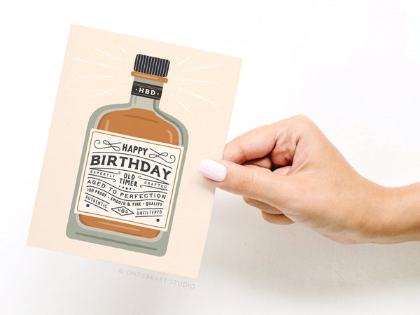 A hand holds the Happy Birthday Whiskey Bottle Greeting Card from Onderkast Studio, featuring a vintage-style illustration with “Old Timer Crafted to Perfection” and festive decorative details.
